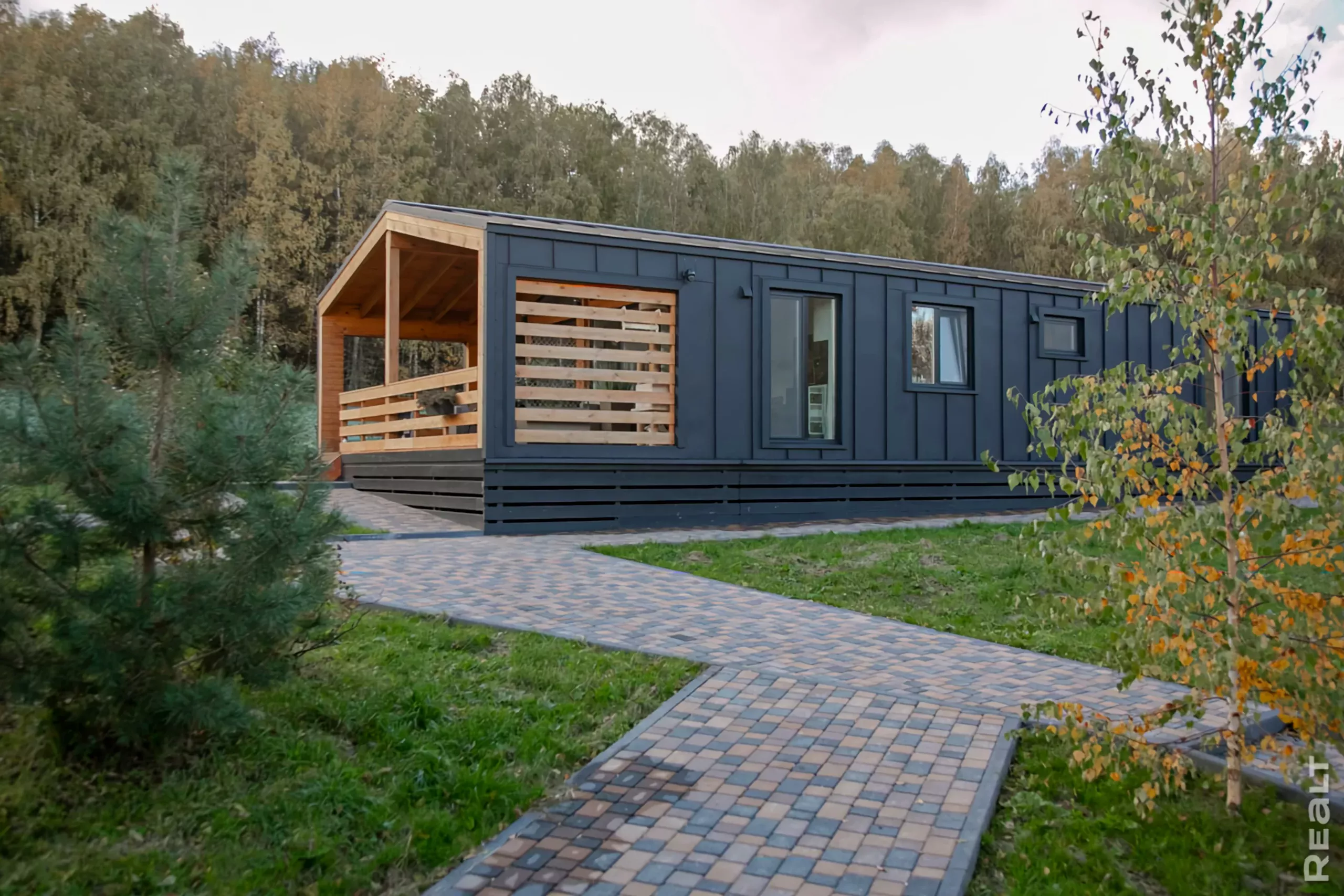 barnhaus-pod-klyuch-proekty-i-ceny-1 Дом барнхаус на фоне красивой зеленой природы с елкой и березкой в переднем плане.